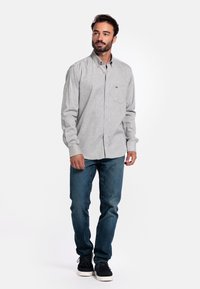Lichtgrijze overhemd met een subtiele textuur, lange mouwen en een enkele borstzak; gecombineerd met donkerblauwe jeans en zwarte schoenen.