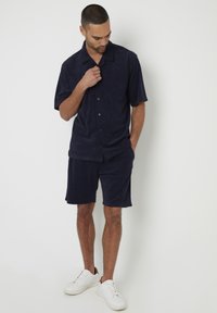 Maglietta blu navy a maniche corte con bottoni, abbinata a pantaloni corti coordinati. Tessuto morbido, design casual, indossata con sneakers bianche.