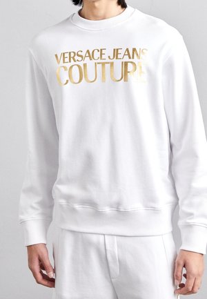 Uomo che indossa una felpa bianca a maniche lunghe e pantaloni, la felpa presenta una scritta dorata in grassetto "VERSACE JEANS COUTURE" sul petto.