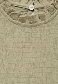 Zeleni teksturirani vrh z ruffled izrezom in majhnim belim gumbom na hrbtni strani. Tkanina ima subtilen mrežast vzorec.