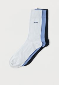 Trois chaussettes d'équipage bien empilées en blanc, bleu et noir avec le texte « BOSS » sur le revers de la chaussette blanche sur un fond uni.