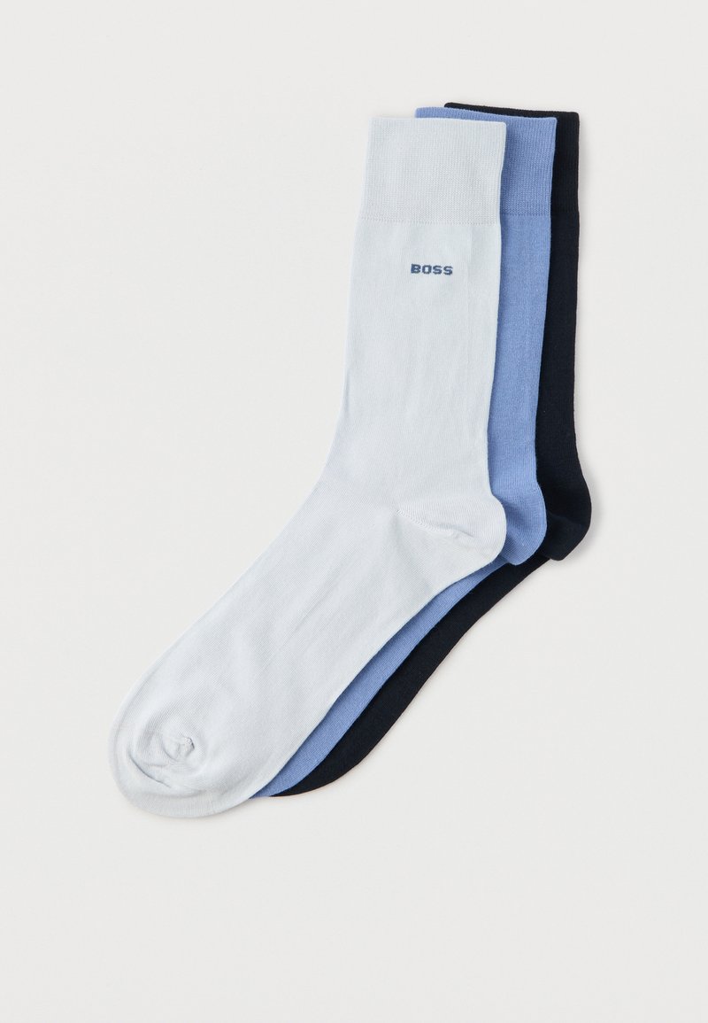 Trois chaussettes d'équipage bien empilées en blanc, bleu et noir avec le texte « BOSS » sur le revers de la chaussette blanche sur un fond uni.