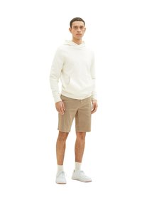 Ein Mann steht in einem cremfarbenen Hoodie, beigen Shorts, weißen Socken und weißen Turnschuhen vor einem schlichten weißen Hintergrund.