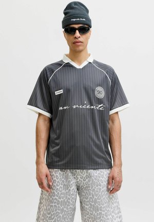 Mann trägt schwarze Mütze, schwarze Sonnenbrille, graues gestreiftes Hemd mit weißem Kragen und hellgraue Leopardenprint-Shorts vor einfachem Hintergrund.