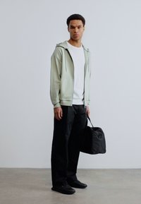 Άνδρας στέκεται σε εσωτερικό χώρο φορώντας ανοιχτό γκρι φούτερ με φερμουάρ, λευκό μπλουζάκι, μαύρο παντελόνι, μαύρα παπούτσια, κρατώντας μια μαύρη τσάντα στο δεξί του χέρι.