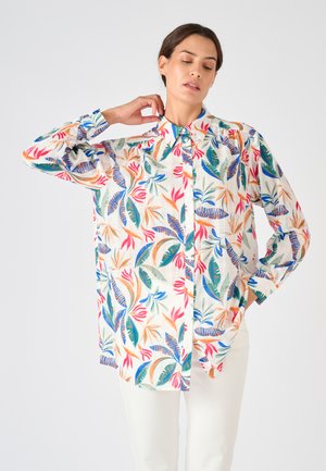 Mujer que lleva una blusa blanca abotonada con estampado colorido de hojas tropicales y flores, posando con una mano en el cuello y la otra en el bolsillo.