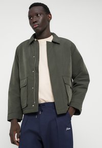 Jachetă ușoară, de culoare verde oliv, cu închidere tip capse, două buzunare mari în față și guler clasic; purtată peste o cămașă bej și pantaloni bleumarin.