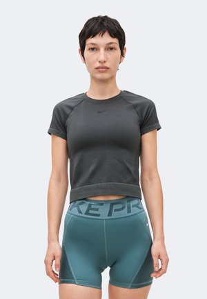 Žena s krátkymi tmavými vlasmi, oblečená v tesnom tmavosivom krátkom tričku a tyrkysových športových šortkách s vysokým pásom a pásom "NIKE PRO".