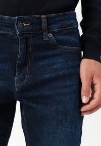 Närbild av mörkblå denimjeans med orange sömmar, knäppt midja, och en persons högra hand vilande vid låret.