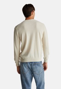 Cremfarbener Pullover mit langen Ärmeln, gerippten Bündchen und Saum. weiche Textur, schlichtes Design. Über blauen Jeans getragen, von hinten betrachtet.