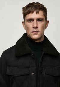 Giacca di suede nera con colletto in shearling verde, chiusura anteriore a pressione, due tasche sul petto e una finitura morbida e testurizzata.