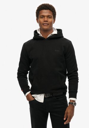 Sudadera negra de tejido suave con un bolsillo frontal, cordones ajustables y puños acanalados, llevada sobre una camiseta blanca y jeans negros.