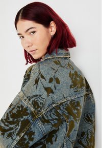 Denim jakna z rožnatim vzorcem v temno zeleni, standardnim ovratnikom in vidnim šivanjem. Tkanina ima rahlo teksturirano površino.