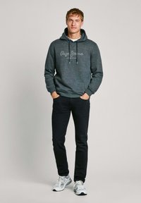 Felpa grigia con cappuccio, cordoncino e logo, abbinata a jeans scuri e sneakers con accenti bianchi. Fit casual e texture liscia.