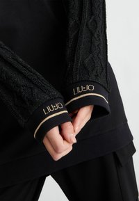 Schwarzer Sweatshirt mit strukturierten, glitzernden schwarzen Bündchen; verfügt über goldene Akzente und ein tailliertes Design. Die Bündchen zeigen den Markennamen in goldener Schrift.