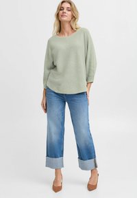 Maglione verde chiaro con texture e scollatura ampia, abbinato a jeans flared in denim blu con risvolti e scarpe con tacco marroni.