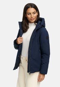 Marineblauwe gewatteerde jasje met capuchon, voorzien van ritssluiting en zijzakken, gedragen over een lichte trui, gecombineerd met beige broek. Textuur van de stof is duidelijk zichtbaar.
