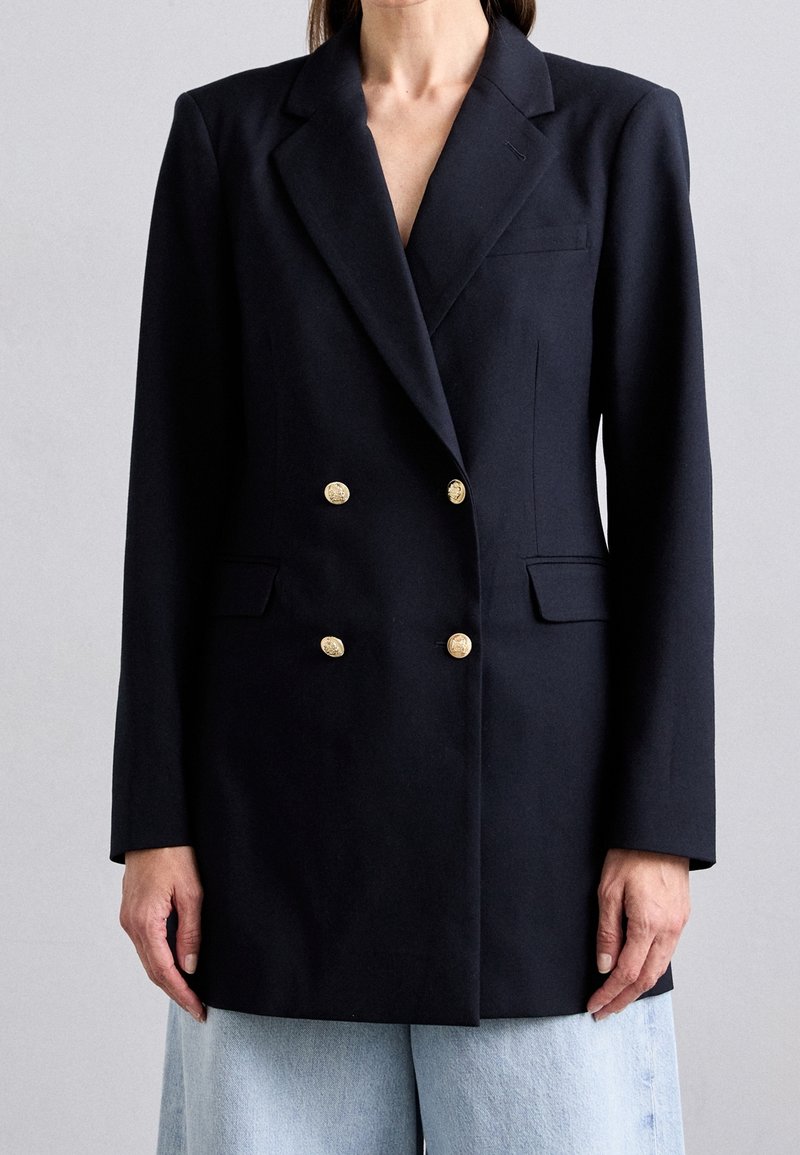 Femme portant un blazer noir croisé avec des boutons dorés et un jean bleu clair, debout devant un fond gris uni.