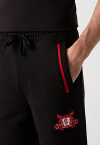 Zwarte sweatpants gemaakt van zachte stof, met een rode gestreepte zak, een elastische tailleband en een opvallend geborduurd logo in rood en wit.
