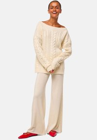 Frau lächelt, trägt einen übergroßen cremfarbenen Strickpullover, weite kremfarbene Hosen und rote spitze Ballerinas, steht vor einem hellen Hintergrund.