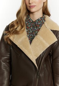 Giacca di pelle marrone con colletto in shearling beige, dotata di tasche con zip e superficie testurizzata. Sotto, una camicia con motivo floreale.