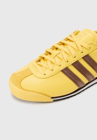 Geltoni odiniai sportbačiai su rudais veliūro dryžiais, tekstūruotu guminio pado ir apvaliu pirštu. Raišteliai geltoni, viršus lygus.