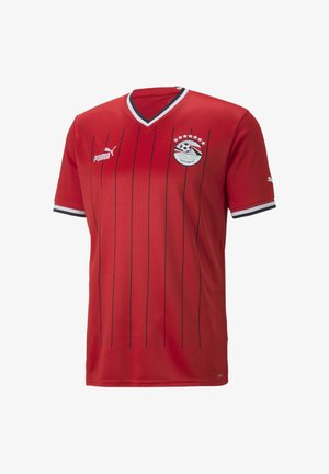 Sarkans futbols krekls ar vertikālām melnām svītrām, ar V veida kaklu, Puma logo kreisajā krūtī un Ēģiptes Futbola asociācijas emblēmu ar zvaigznēm.