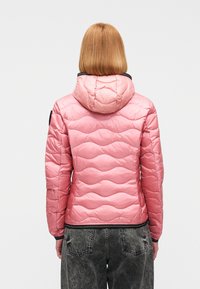 Άτομο με ίσια, ξανθά μαλλιά μήκους μέχρι τους ώμους, που φοράει ροζ καπιτονέ μπουφάν με κουκούλα και ξεθωριασμένα μαύρα τζιν, φανερό από πίσω.