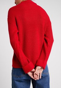 Röd stickad tröja med en strukturerad ribbdesign, vida ärmar och en lös passform. Har hög krage och manschetter på ärmarna.