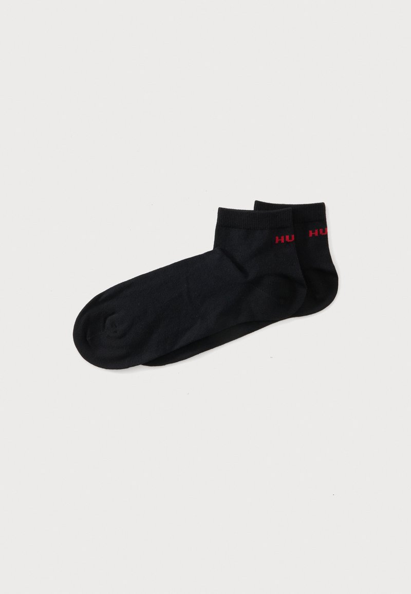Paire de chaussettes noires aux chevilles avec un texte partiel rouge sur les bord-côtes, posées à plat sur un fond blanc.