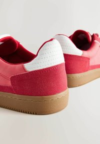 Rote Turnschuhe verfügen über ein glattes Leder- und strukturiertes Wildlederdesign, mit einer weißen gepolsterten Ferse und einer Gummisohle, die horizontale Rillen aufweist.