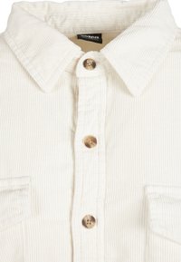 Chemise en velours côtelé écru avec col pointu, devant boutonné avec trois boutons marron, et vue partielle de deux poches poitrine.