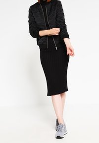 Kobieta w czarnej pikowanej kurtce i prążkowanej spódnicy midi w połączeniu z szarymi sneakersami, poprawiająca rękaw na białym tle.