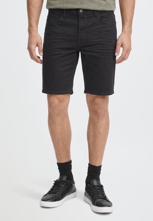 Modèle portant un short en jean noir jusqu’aux genoux, des chaussettes noires et des baskets noires à semelles blanches, sur un fond uni.