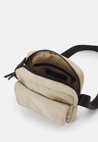 Borsa in tela beige con manico nero, dotata di scomparto principale con zip e tasca frontale. Texture liscia, forma rettangolare, dettagli minimal.