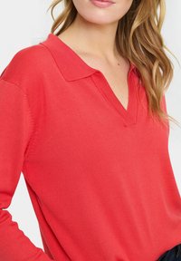 Korálový sveter s výstrihom do V a golierom, vyrobený z rebrovanej tkaniny. Obsahuje dlhé rukávy, hladkú textúru a priliehavý strih.