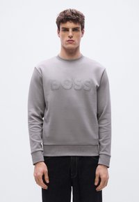 Γκρι φούτερ με υφή λογότυπου "BOSS" στο μπροστινό μέρος, σχέδιο με στρογγυλή λαιμόκοψη, ριμπ μανσέτες και άνετη εφαρμογή. Φορεμένο με σκούρο τζιν.