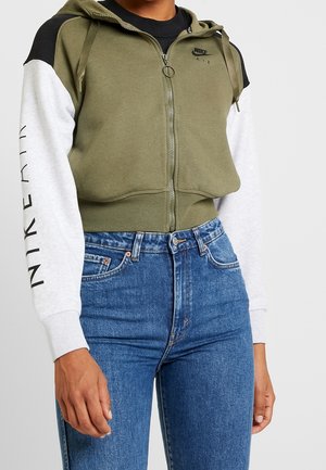 Person wearing olive green zip-up Nike Air hoodie with white and black sleeves and blue high-waisted jeans, standing against white background.