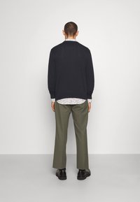 Námořnický cardigan přes bílou vzorovanou košili, olivově zelené kalhoty a černé boty. Jednoduchý design a volný střih viditelné zezadu.