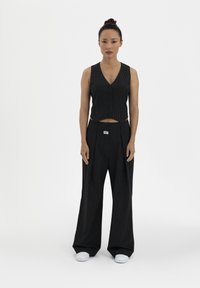 Black pinstriped vest with a V-neck, button front, and cropped design, paired with loose-fitting pinstriped trousers, worn with white sneakers.