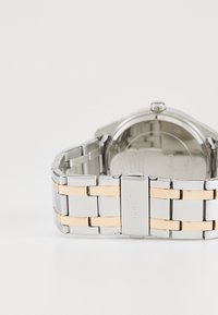 Montre-bracelet en acier inoxydable, de couleur argent et or, avec des maillons rectangulaires et une finition polie. Elle présente un boîtier rond et un cadran simple.