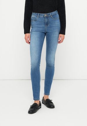 Vrouw draagt lichtblauwe skinny jeans, zwarte zachte trui en zwarte leren loafers, staand tegen een witte achtergrond.