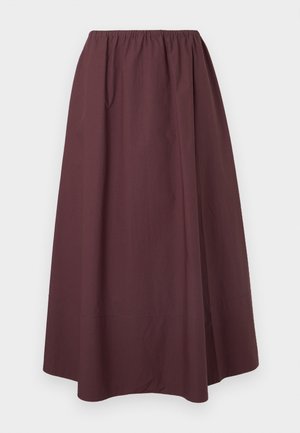 Vrouw met lang zwart haar en bril, die een lichtgroen oversized shirt draagt over een donkerbruin topje en een lange rok, staand op een grijze vloer.