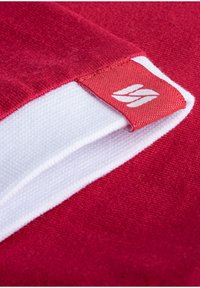 Roter Baumwollstoff mit einem weißen Akzent am Saum. Verfügt über ein kleines rotes Etikett mit einem weißen Logo. Glatte Textur mit einem sauberen Finish.