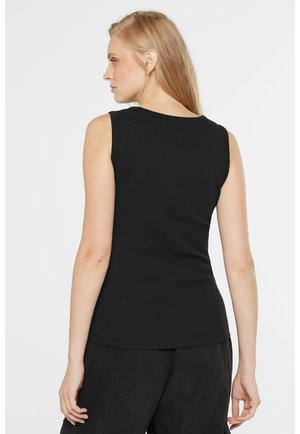 Blonde Frau, von hinten gesehen, trägt ein ärmelloses, eng anliegendes schwarzes Oberteil und lockere schwarze Hose vor weißem Hintergrund.