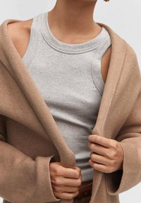 Persona vestida con una camiseta de tirantes acanalada gris claro debajo de un abrigo beige, sosteniendo el abrigo abierto a nivel del pecho con ambas manos.