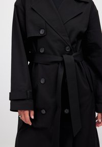 Čierny trench coat s dvojradovým zapínaním, veľkými klopami a pásom v páse. Obsahuje čierne gombíky a hladkú textúru.