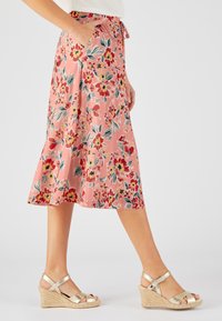 Falda midi floral en rosa, con cintura ajustable, bolsillos laterales y un diseño fluido. Sandalias de cuña con acabado metálico.