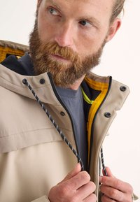 Un homme barbu portant une veste beige avec une doublure jaune, tirant la fermeture éclair d'une main et regardant sur le côté, contre un fond neutre.