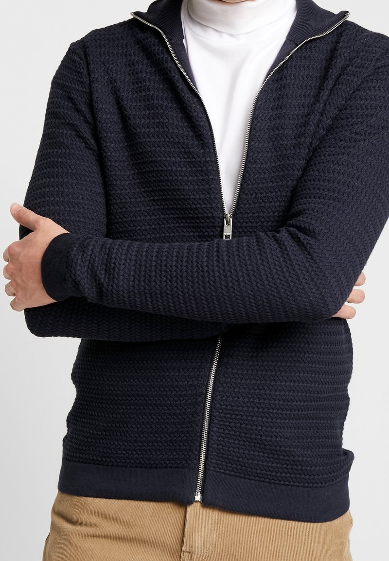 Homme portant un pull zippé bleu marine texturé par-dessus un col roulé blanc, les bras croisés, et un pantalon en velours côtelé beige visible de la taille vers le bas.